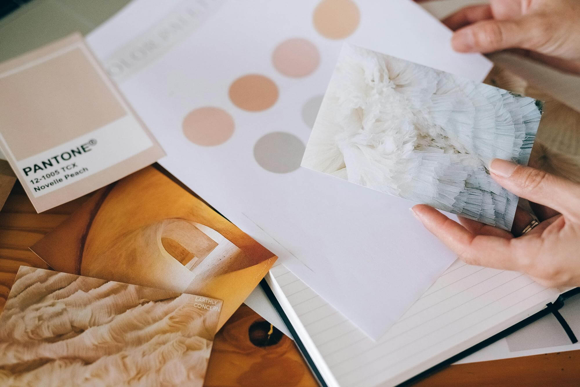 A persons hands hold textured photos over a desk with a Pantone color swatch labeled 12-0910 TCX Novelle Peach and color samples in shades of beige and peach. A notebook and design materials are also on the desk.