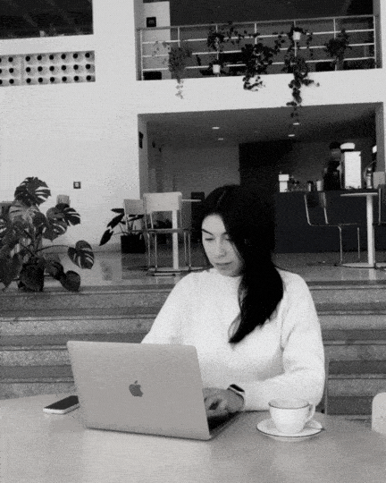 A woman sits at a table working on a laptop in a modern, open indoor space. A cup and saucer are on the table beside her.