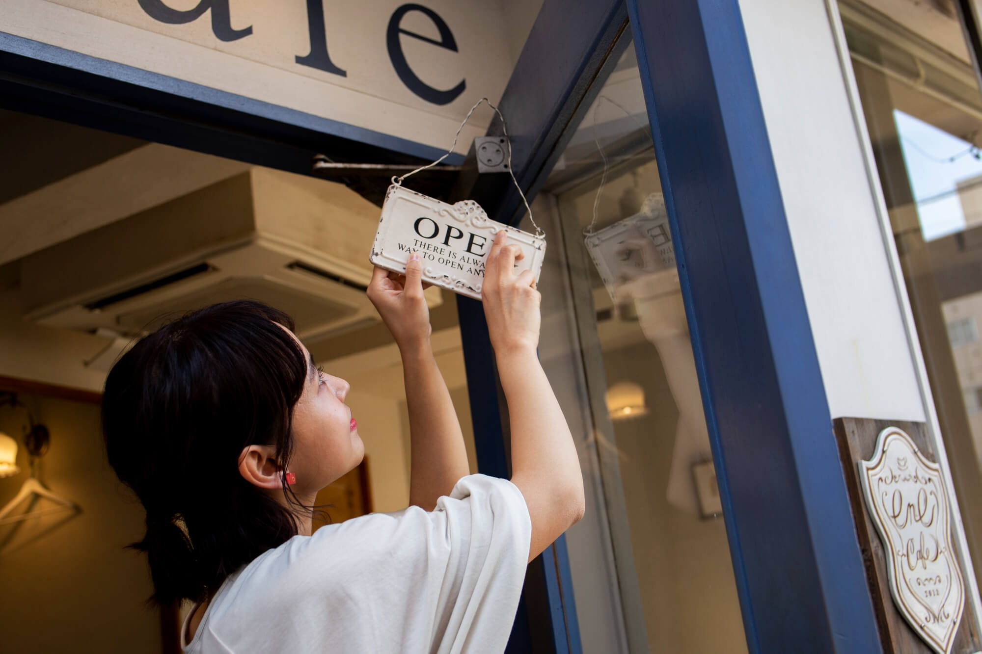 A person wearing a white shirt adjusts an OPEN sign at the entrance of a café. The café has a blue door frame and warm lighting inside.