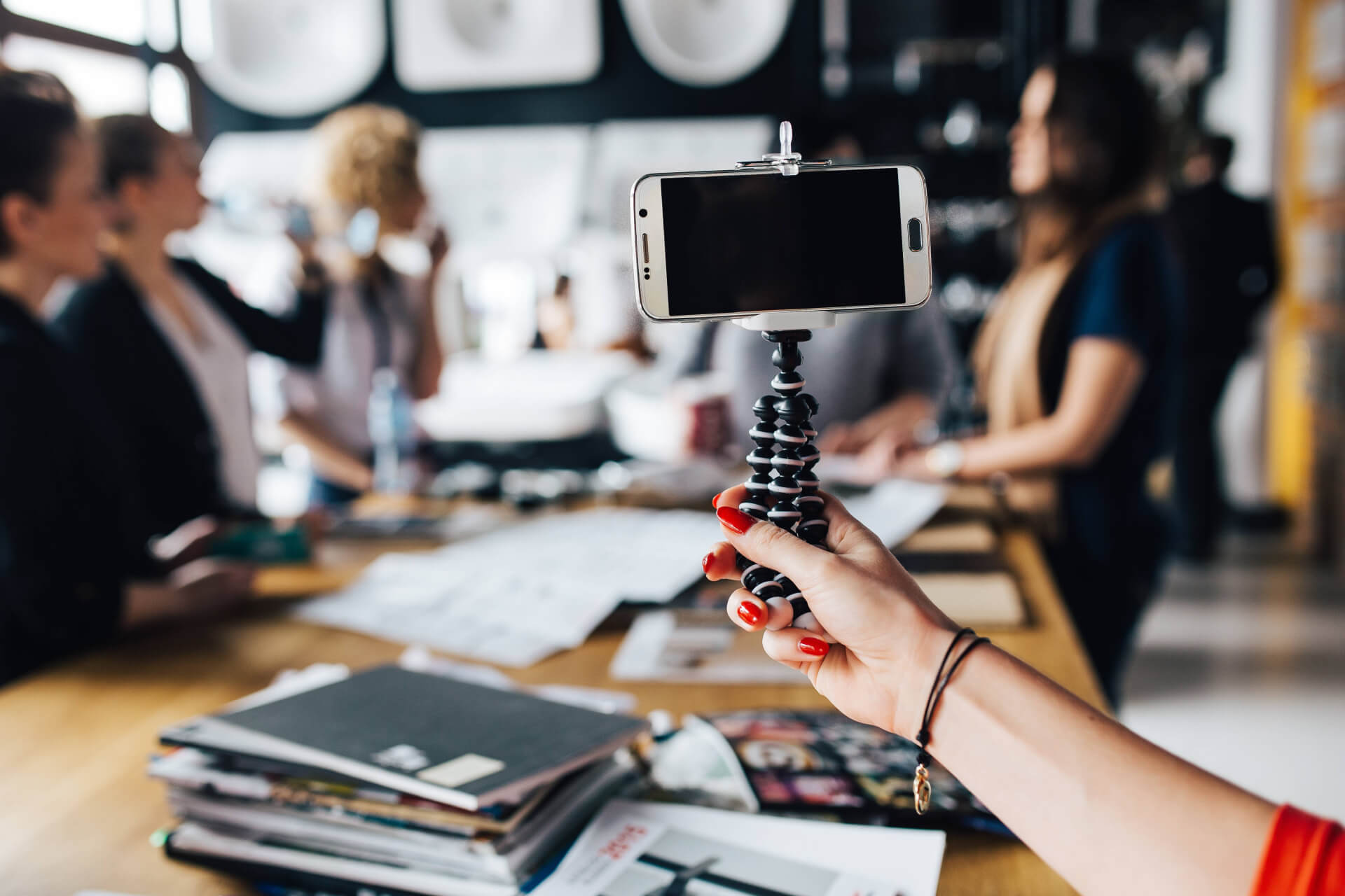 A person holds a smartphone on a flexible tripod, recording a group of people having a discussion around a table filled with papers and magazines. The focus is on the smartphone, while the people are blurred in the background.